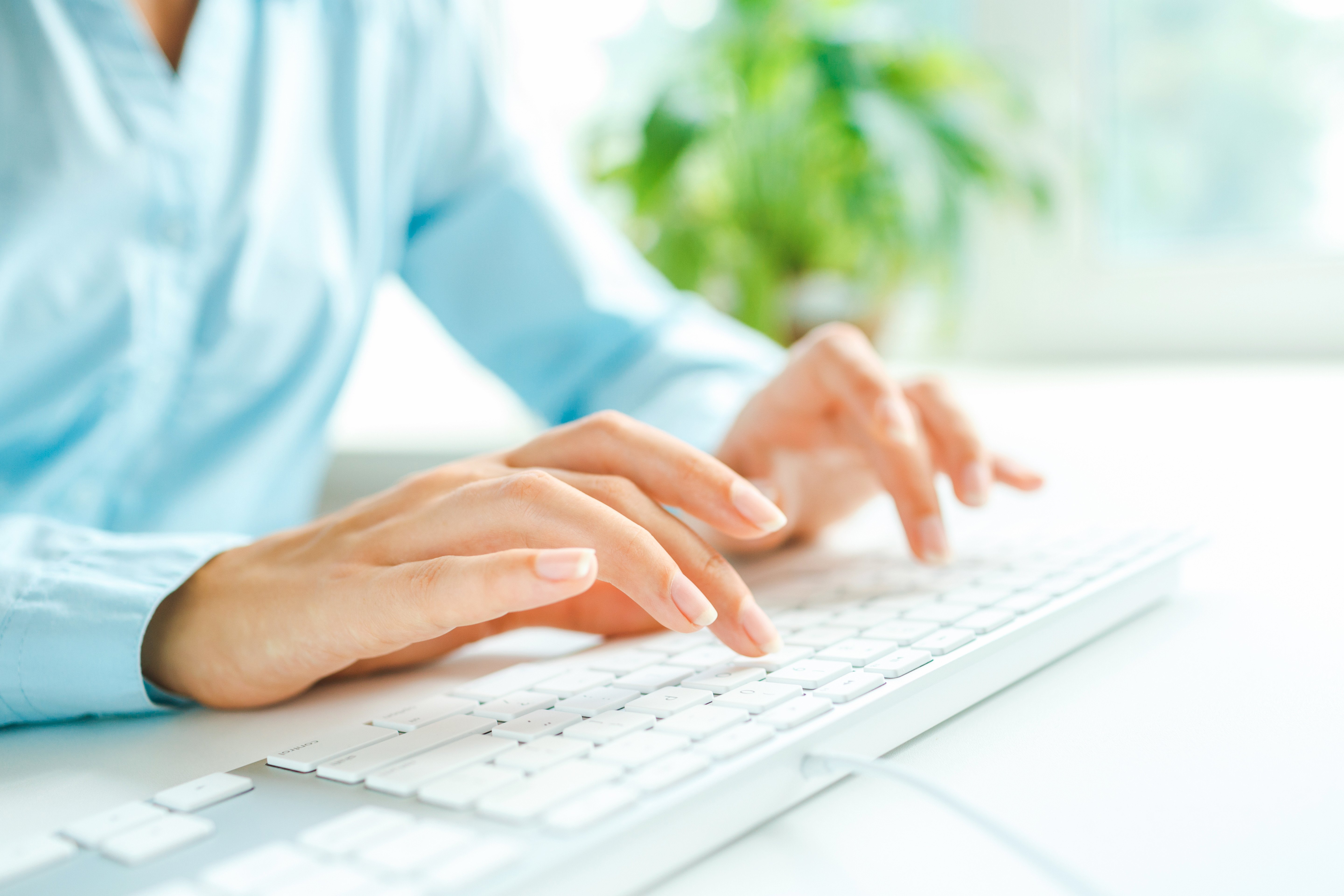 The image shows a close-up of a person typing on a white keyboard. The individual is wearing a light blue shirt, and the background features a blurred green plant, giving the scene a clean and professional atmosphere.