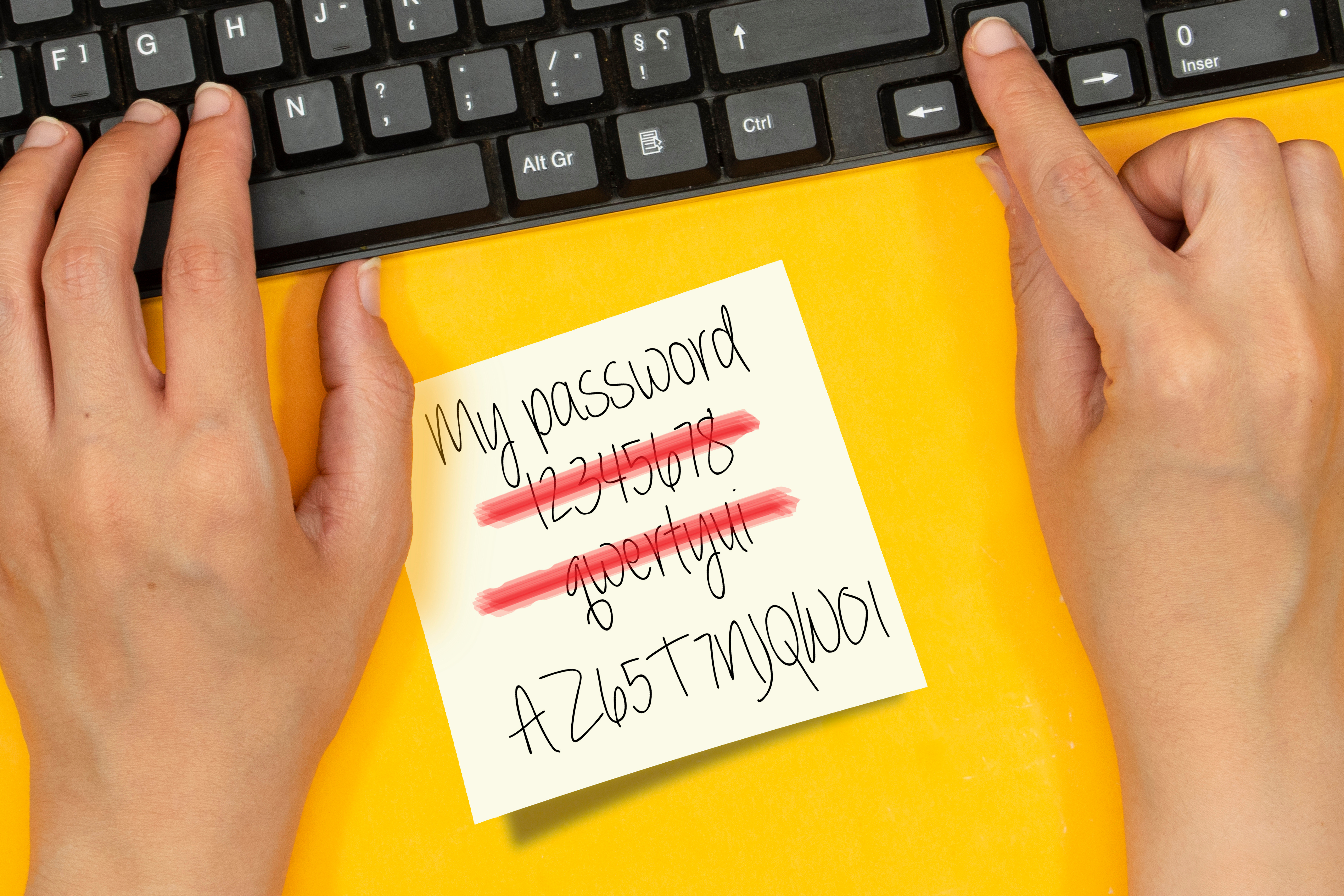 Hands typing on a keyboard, with a sticky note on a yellow background with three passwords written on it. Two are scratched out and one remains legible.